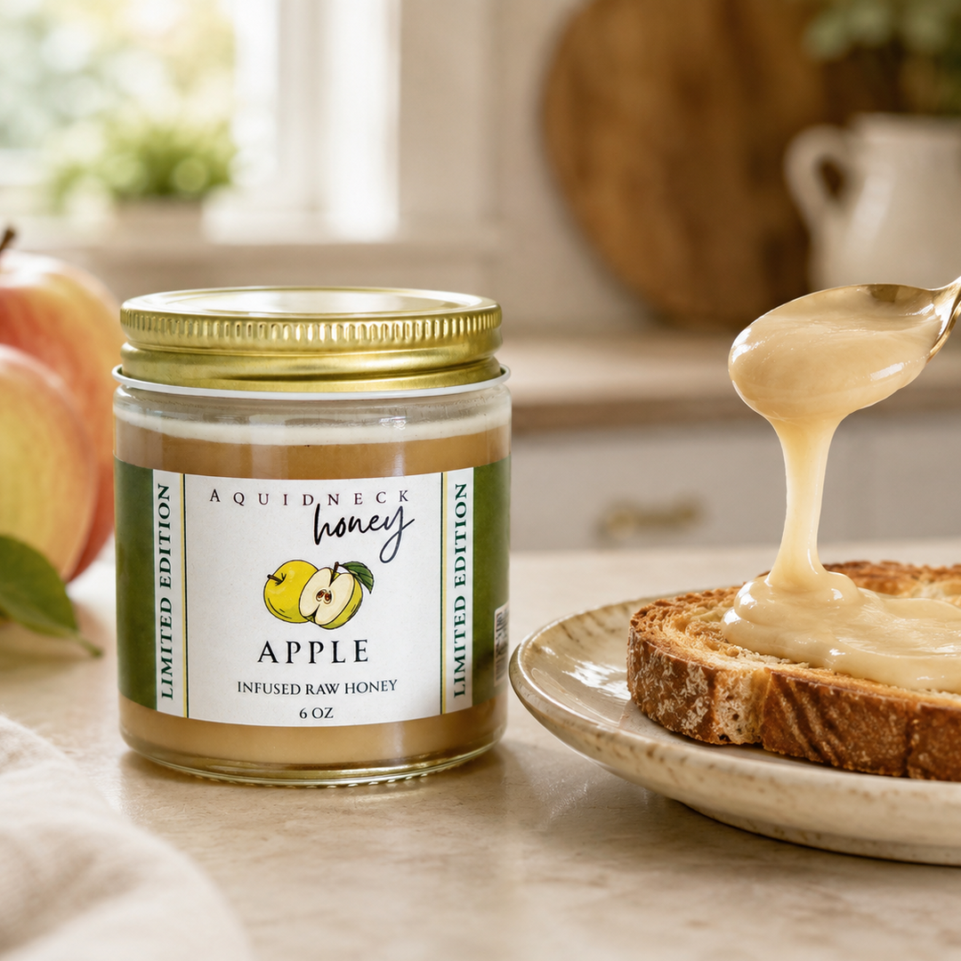 A jar of Aquidneck Honey Apple Infused Raw Honey sits on a kitchen counter next to a slice of toast with honey being drizzled from a gold spoon.